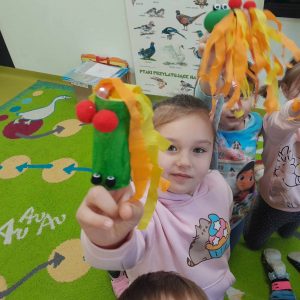 Uczennica oddziału przedszkolnego widoczna na pierwszym planie trzyma w wyciągniętej przed siebie dłoni dmuchawkę przypominającą smoka wawelskiego z długim ogonem wykonanym z krepy. Dziewczynka ma włożone dwa palce wewnątrz dmuchawki. W tle widać częściowi dwoje uczniów, którzy również trzymają w wyciągniętych dłoniach swoje dmuchawki.