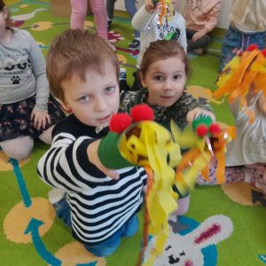 Uczniowie oddziału przedszkolnego siedzą na dywanie. Dzieci trzymają w dłoniach wykonane przez siebie dmuchawki przypominające smoka wawelskiego. Dwoje dzieci na pierwszym planie wyciąga przed siebie swoje dmuchawki.