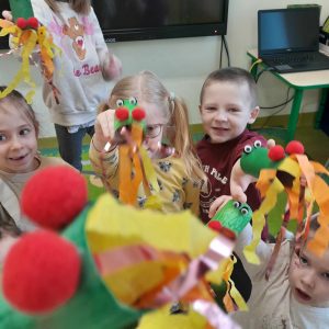 Uczniowie oddziału przedszkolnego siedzą na dywanie. Dzieci trzymają w wyciągniętych przed siebie dłoniach wykonane przez siebie dmuchawki przypominające smoka wawelskiego.