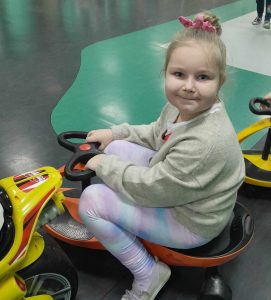 Uczennica siedzi na pomarańczowym pojeździe z kierownicą. Dziewczynka uśmiecha się do aparatu.