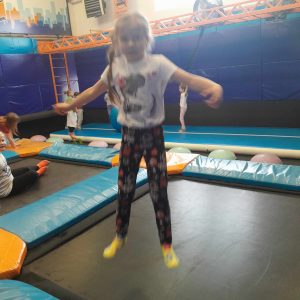 Uczennica oddziału przedszkolnego podskakuje na trampolinie. Dziewczynka ma szeroko rozstawione ramiona.