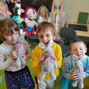Troje uczniów oddziału przedszkolnego stoją w rzędzie. Dzieci tulą pluszowe króliczki. W tle widać ułożone na krzesłach wielkie kukły - marzanny.