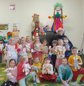 Uczniowie oddziału przedszkolnego pozują do wspólnego zdjęcia wraz z Panią Dyrektor. Dzieci przytulają pluszowe króliczki. W tle widać dwie duże kukły - marzanny.