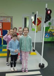 Pięć uczennic oddziału przedszkolnego stoi w parach przed przejściem dla pieszych w miasteczku ruchu drogowego. Obok nich na słupku umieszczona jest sygnalizacja wskazująca czerwone światło.