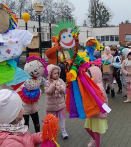 Uczniowie oddziału przedszkolnego stoją na terenie szkoły trzymając w wyciągniętych w górę dłoniach wielkie lalki przypominające marzanny.
