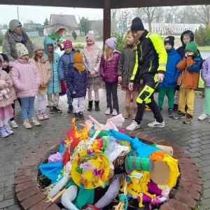 Uczniowie stoją w kręgu pp środku którego ułożono na siebie przyniesione przez uczniów marzanny. Między kręgiem a uczniami przechodzi Pan przyglądający się ułożonemu stosowi.