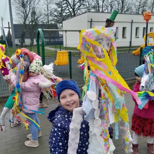 Uczniowie oddziału przedszkolnego stoją na terenie szkoły trzymając w wyciągniętych w górę dłoniach wielkie lalki przypominające marzanny. Dziewczynka widoczna na pierwszym planie szeroko uśmiecha się trzymając w dłoniach dużą kukłę.