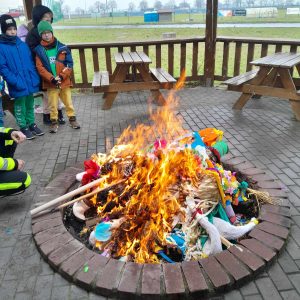 Zdjęcie ogniska, w którym palą się marzanny. W tle widoczne są drewniane ławki oraz stojący uczniowie patrzący na ognisko.