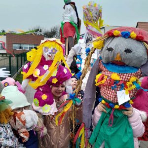 Uczniowie oddziału przedszkolnego stoją na terenie szkoły trzymając w wyciągniętych w górę dłoniach wielkie lalki przypominające marzanny.