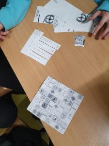 Zdjęcie stołu na którym leży kartka z czarno-białą mozaiką oraz ułożone paski papieru z napisami tworzące wskazówkę.