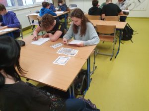 Uczniowie klasy ósmej siedzą w grupach przy połączonych stołach. Na pierwszym planie widać stół z rozłożonymi elementami zadania nad którymi pochyla się dwoje uczniów.