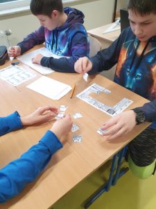 Dwóch uczniów siedzi obok siebie przy stole. Jeden z nich układa mapę z puzzli, a drugi wykonuje obliczenia na kartce.