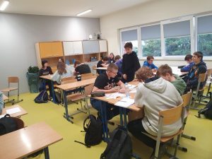 Uczniowie klasy ósmej siedzą w sali matematycznej przy rozstawionych stołach do pracy w grupach. Jeden z uczniów stoi.