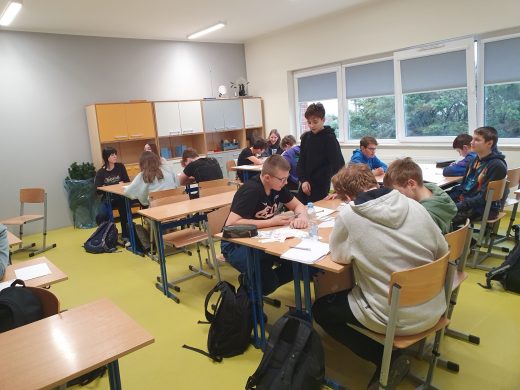 Uczniowie klasy ósmej siedzą w sali matematycznej przy rozstawionych stołach do pracy w grupach. Jeden z uczniów stoi.