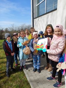 Uczniowie klasy drugiej pozują do wspólnego zdjęcia przy ścianie szkoły. Dzieci trzymają w dłoniach znalezione prezenty oraz rysunek wielkanocnego zajączka z kolorowymi pisankami.