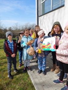 Uczniowie klasy drugiej pozują do wspólnego zdjęcia przy ścianie szkoły. Dzieci trzymają w dłoniach znalezione prezenty oraz rysunek wielkanocnego zajączka z kolorowymi pisankami.