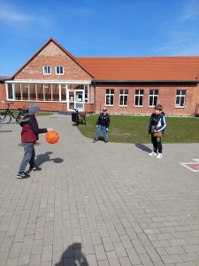 Trzech uczniów klasy drugiej gra piłką do koszykówki na szkolnym boisku. Jeden z chłopców kozłuje piłką.