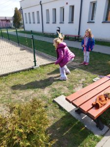 Uczennica klasy drugiej przechodzi przez gumę do skakania zapieczoną o ławkę i płotek na terenie przed szkołą.