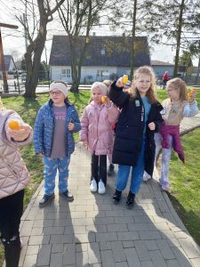 Uczennice klasy drugiej stoją na chodniku przy szkole. Dziewczynki trzymają w dłoniach żółte plastikowe jajeczka.