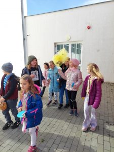 Uczniowie klasy drugiej stoją przed budynkiem szkoły. Grupka dzieci trzyma na wysokości swoich głów żółty półprzezroczysty worek.
