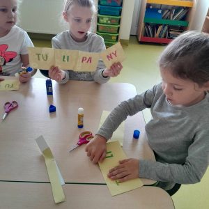 Uczennica siedzi przy stole i skleja dwie karteczki z literami. Obok jej koleżanka trzyma w dłoniach karteczki z literami tworzące napis: 'TULIPANY".