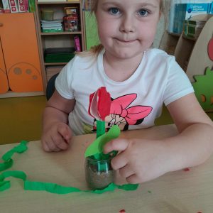 Uczennica oddziału przedszkolnego siedzi przy stoliku. Przed nią stoi szklana doniczka w którą włożony jest papierowy kwiatek ozdabiany przez dziewczynkę zieloną krepą.