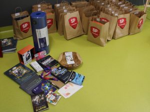 Zdjęcie ułożonych na podłodze nagród, które otrzymali laureaci konkursu. Obok wyłożonych nagród stoją zapakowane szare torby z logo szkoły.