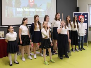 Szkolny chórek podczas występu na gali. Uczniowie obrani są w białe bluzki i granatowe spódnice lub spodnie. Stoją w dwóch rzędach. W pierwszym rzędzie stoją dwie uczennice trzymające mikrofon.