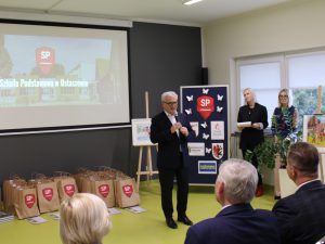 Zdjęcie Starosty Toruńskiego wykonane podczas przemowy na gali wręczenia nagród. Pan Starosta trzyma mikrofon w dłoni. Za nim widoczne są ustawione na podłodze szare torby z nagrodami. Po prawej stronie widać również tablicę z logo szkoły i sponsorów konkursu obok której stoją dwie nauczycielki.