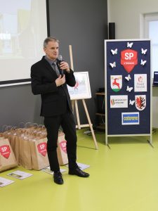 Zdjęcie wykonane podczas przemowy przedstawiciela Kuratorium Oświaty. Pan trzyma w dłoniach mikrofon. Za nim na podłodze leżą torby z nagrodami dla laureatów konkursu a po prawej stronie widoczna jest tablica z logo szkoły oraz patronów konkursu.