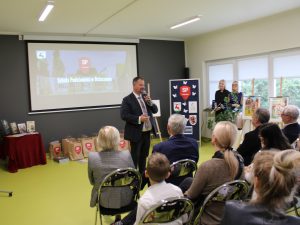 Zdjęcie wykonane podczas przemowy Przewodniczącego Rady Gminy Łysomice. Przewodniczący trzyma w dłoniach mikrofon. Za nim na podłodze leżą torby z nagrodami dla laureatów konkursu a po prawej stronie widoczna jest tablica z logo szkoły oraz patronów konkursu. Przed mówcą widać siedzących na krzesłach uczestników gali.