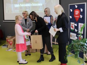 Uczestniczka konkursu odbiera torbę z nagrodą. Dziewczynka ściska dłoń przewodniczącej jury a obok stoją Pani Dyrektor oraz dwie nauczycielki. W tle po prawej stronie widoczna jest tablica z logo szkoły i patronami konkursu.
