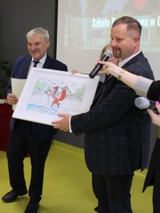 Wójt Gminy Łysomice oraz Przewodniczący Rady Gminy wspólnie trzymają oprawioną w białą ramę kopię wyróżnionej w konkursie pracy. Pan Przewodniczący mówi do mikrofonu.