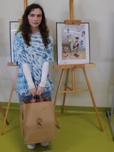 Uczennica nagrodzona w konkursie pozuje obok swojej oprawionej i ustawionej na drewnianej sztaludze pracy. Dziewczynka trzyma w dłoniach przed sobą torbę z nagrodami.