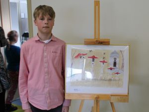 Uczeń pozuje do zdjęcia obok wyróżnionej w konkursie swojej pracy. Praca została oprawiona w ramkę i ustawiona na drewnianej sztaludze.