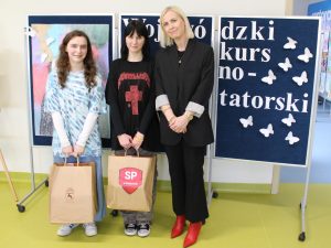 Dwie nagrodzone w konkursie uczennice pozują do wspólnego zdjęcia z nauczycielką. Dziewczynki trzymają w dłoniach torby z nagrodami. W tle widoczna jest niebieska tablica z białym napisem "Wojewódzki konkurs plastyczno-recytatorski".