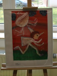 Zdjęcie ustawionej na drewnianej sztaludze oprawionej w białą wąską ramkę pracy wyróżnionej w konkursie. Praca przedstawia Pinokia uciekającego przed statkiem kosmicznym.