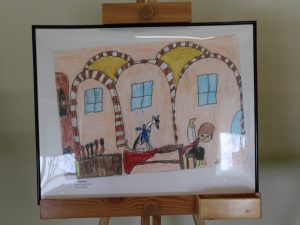 Zdjęcie ustawionej na drewnianej sztaludze oprawionej w czarną wąską ramkę pracy wyróżnionej w konkursie. Praca przedstawia chłopca szyjącego buty, któremu przygląda się pies.