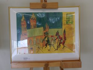 Zdjęcie ustawionej na drewnianej sztaludze oprawionej w złotą wąską ramkę pracy wyróżnionej w konkursie. Praca przedstawia Pinokia idącego w towarzystwie dwóch lisów przez miasto.