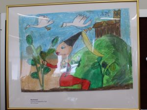 Zdjęcie ustawionej na drewnianej sztaludze oprawionej w złotą wąską ramkę pracy wyróżnionej w konkursie. Praca przedstawia księżniczkę nad którą lecą dwa łabędzie.