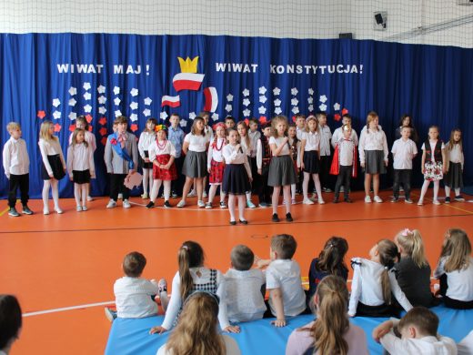 Uczniowie występujący na apelu stoją razem na środku sali gimanstycznej. Przed nimi stoją dwie dziewczynki z mikrofonami. W tle na niebieskim materiale zawieszona została dekoracja - napis "WIWAT MAJ! WIWAT KONSTYTUCJA!" a pod nim biało czerwone kwiatki, trzy flagi Polski oraz korona tworzące razem wizerunek orła.