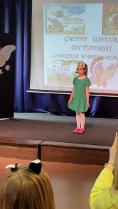 Uczennica w zielonej sukience stoi na scenie. Dziewczynka trzyma dłońmi sukienkę na brzegu. Przy ustach ma mikrofon a na głowie żółty kwiatek. W tle wyświetla się napis "GMINNY KONKURS RECYTATORSKI "ZWIERZĘTA W POEZJI DZIECIĘCEJ"".