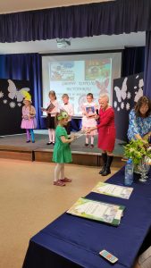 Uczennica w zielonej sukience odbiera z rąk Pani nagrodę. W tle na scenie stoi czworo dzieci.