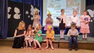 Uczniowie nagrodzeni w konkursie stoją lub siedzą na scenie. Dzieci trzymają książki. Jedna z dziewczynek, która siedzi pokazuje swojej koleżance książkę.