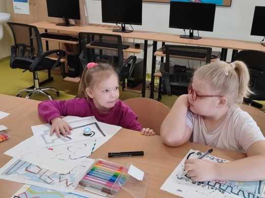 Dwie uczennice klasy pierwszej siedzą przy dużym stole. Dziewczynki patrzą na siebie. Na stole przed nimi leżą kartki z rysunkami, pisaki i świecąca kulka. Jedna z dziewczynek trzyma w dłoni pisak.