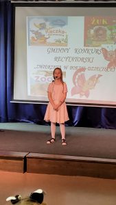 Uczennica w jasnej sukience stoi na scenie. Dziewczynka ma splecione przed sobą dłonie a przy buzi mikrofon przenośny. W tle wyświetla się na rzutniki napis "GMINNY KONKURS RECYTATORSKI "ZWIERZĘTA W POEZJI DZIECIĘCEJ"". Wokół napisu umieszczone są kolorowe grafiki.