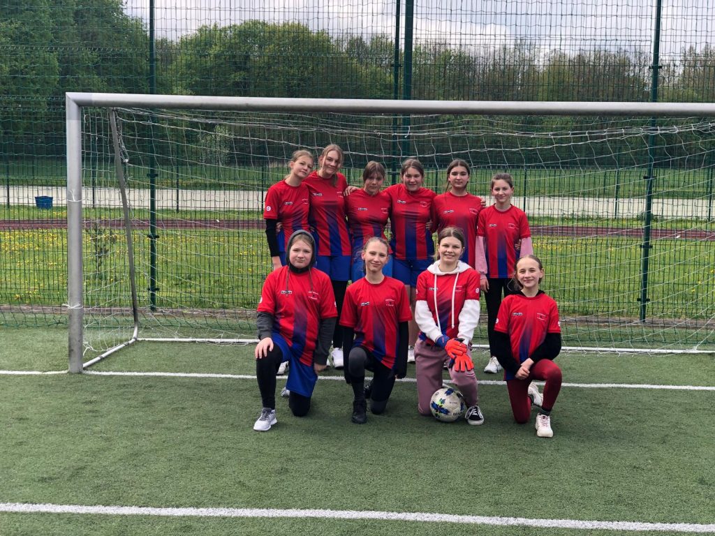 Mistrzynie Gminy Łysomice w piłce nożnej pozują do wspólnego zdjęcia na orliku (ID). Część uczennic stoi w świetle bramki a przed nimi cztery klękają na jednym kolanie.