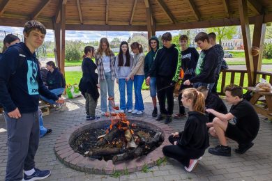 Uczniowie klasy ósmej stoją wokół ogniska, nad którym trzymają kijki z nadzianymi na nich kiełbaskami. Część uczniów dla wygody kuca przy ognisku.