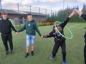 Trzech chłopców stoi na murawie boiska i trzyma się za ręce. Jeden z nich pochyla się i przekłada przez wyciągniętą ręką i głowę hula hop.