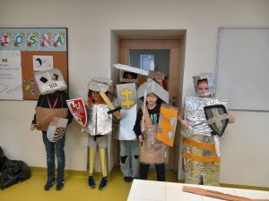 Pięcioro uczniów pozuje do wspólnego zdjęcia w wykonanych przez siebie zbrojach rycerskich. Chłopcy trzymają tarcze i miecze, a na głowach mają hełmy.